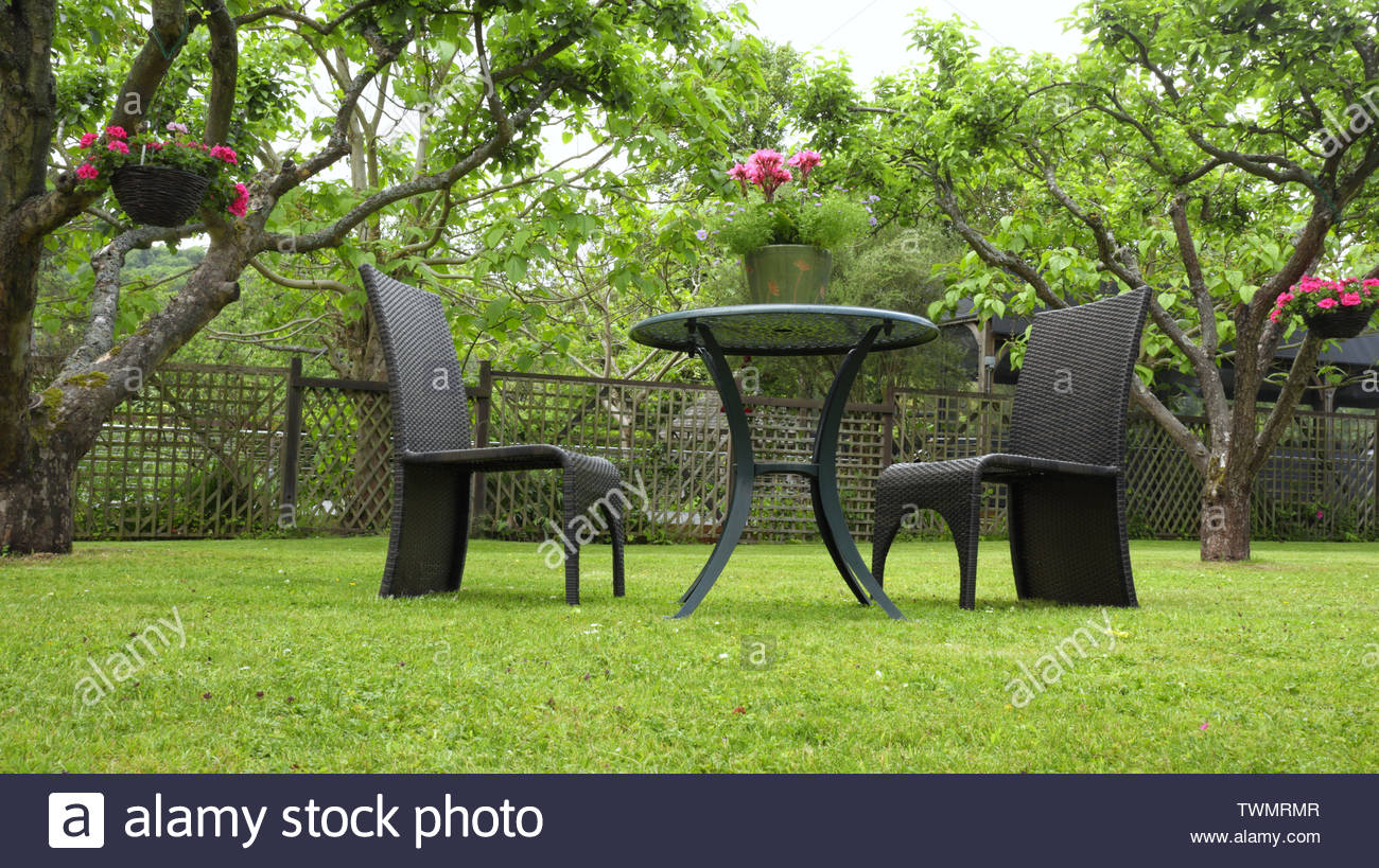 deux chaises en rotin et table avec pot de fleurs sur l herbe dans un jardin d ete entouree par des arbres des paniers suspendus colores twmrmr