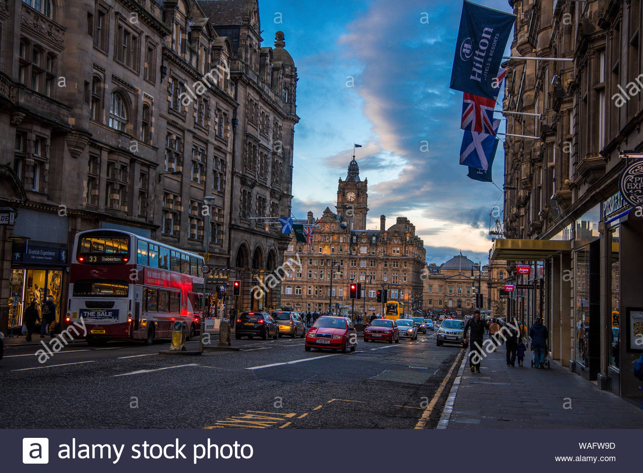 moody street scene of the royal mile edinburgh scotland uk showing hilton hotel old tall buildings shops cars buses and people plus flags WAFW9D